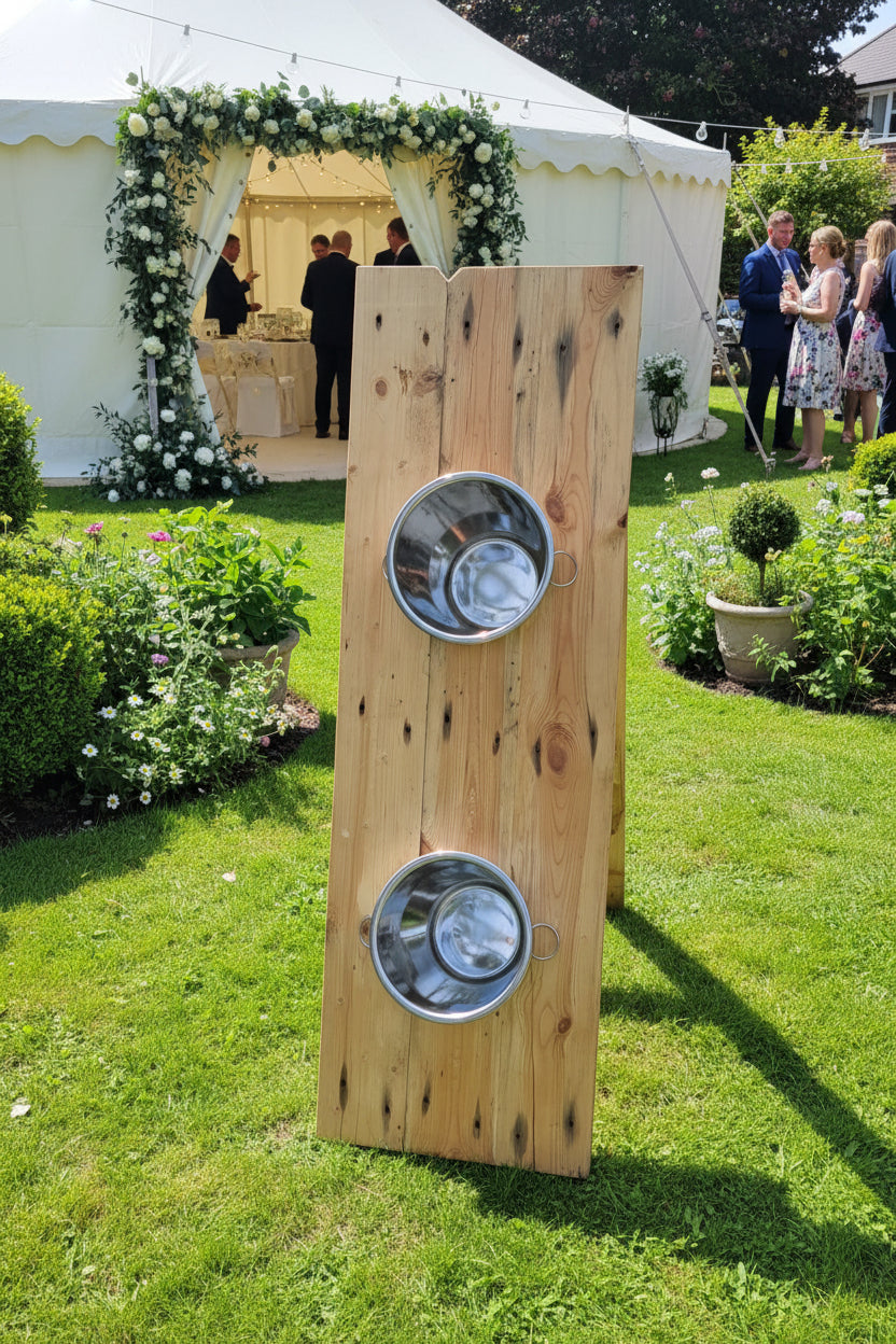 Upright wooden ball in a bucket game with two metal wine buckets, set against a marquee in a garden background.