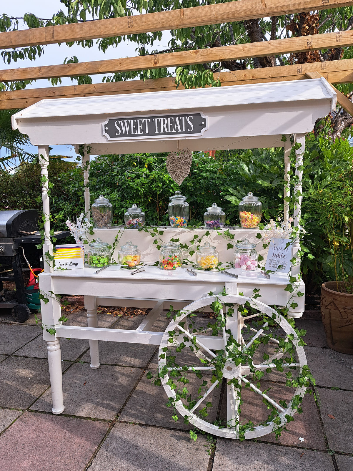 Decorative sweets cart with flowers and a sign on a stone patio in a private garden
