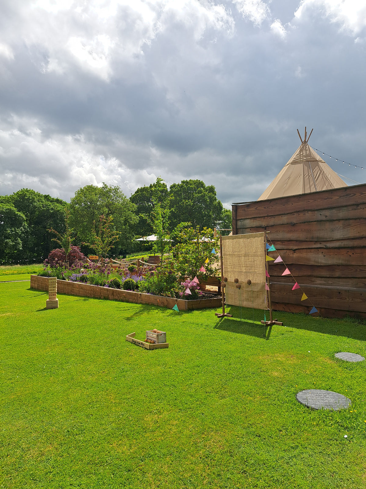 Outdoor coconut shy game setup with hessian and colourful flags on a grassy area with a tipi and trees in the background.