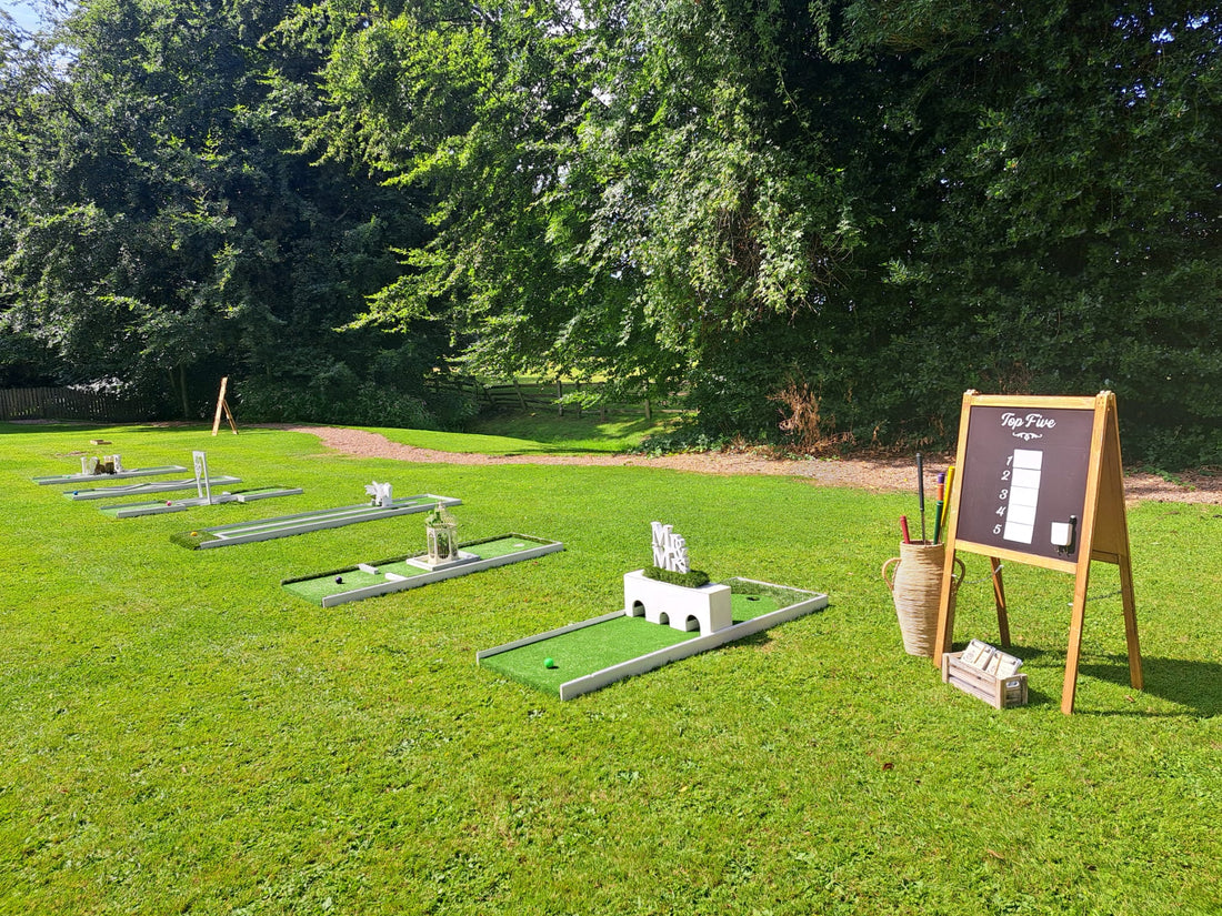 Outdoor mini golf course setup on a grassy lawn with several holes with decorative white obstacles, a wooden sign, and a tall basket holding clubs, surrounded by dense trees. Mitton Hall, Clitheroe