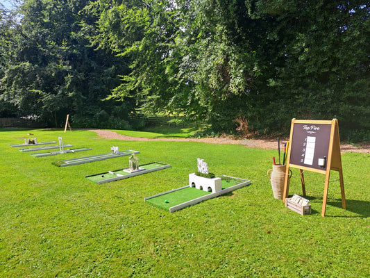 Outdoor mini golf course setup on a grassy lawn with several holes with decorative white obstacles, a wooden sign, and a tall basket holding clubs, surrounded by dense trees. Mitton Hall, Clitheroe