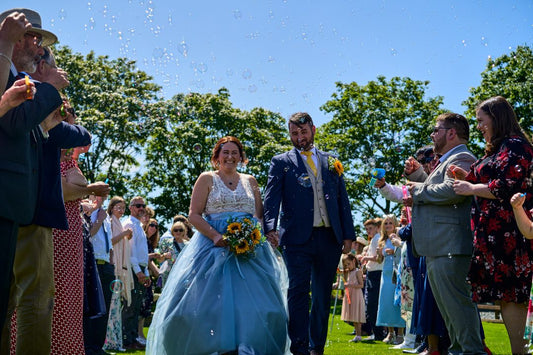 This image shows Sian & Adam, the companys owner, on their wedding day, they are surrounded by guests blowing bubbles as the walk through an isle on grass with blue skies.