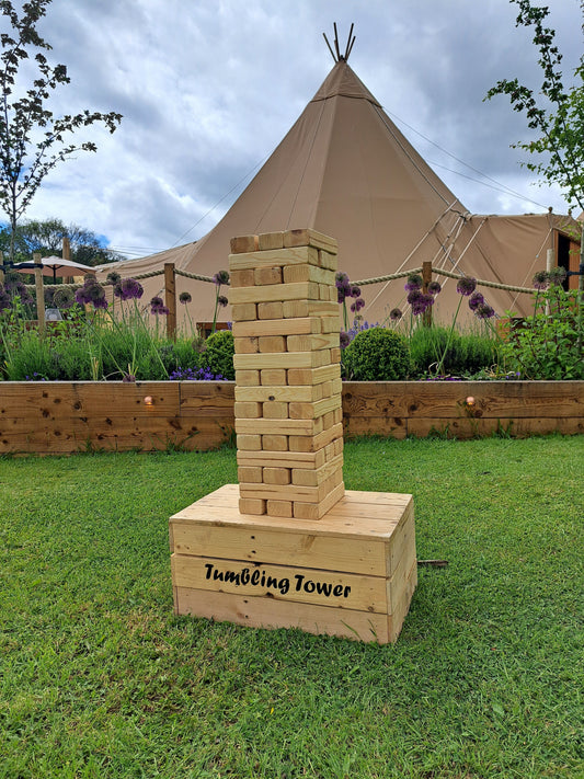 A giant tumbling tower game on grass with a flower boarder and a beige wedding tipi set up in the background