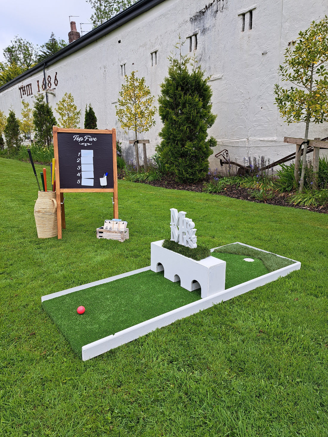 A wedding golf hole with a white box as an obstacle and a scoreboard with a basket of golf clubs, set up on a lawn and a white building in the background