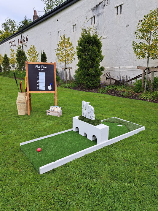 A wedding golf hole with a white box as an obstacle and a scoreboard with a basket of golf clubs, set up on a lawn and a white building in the background