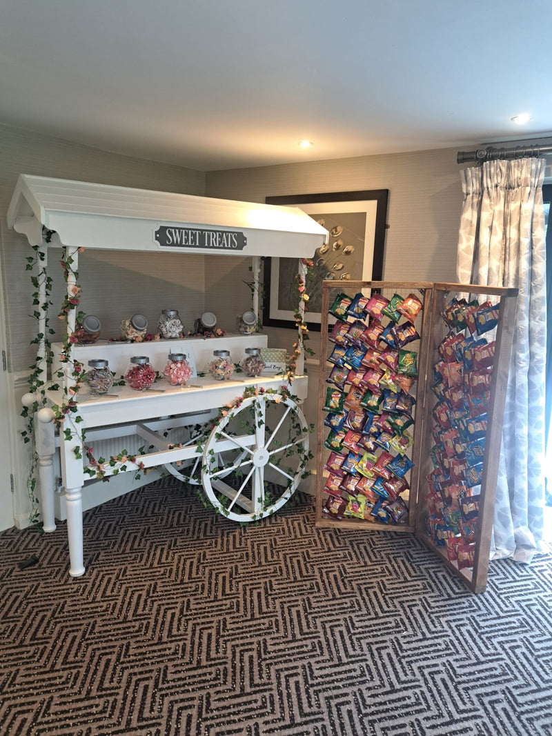 A white decorative cart styled like a vintage candy stand, with shelves holding small jars and bags, a hanging sign reading 'SWEET TREATS,' and a wooden folding screens covered in colourful packaged snacks. The setup is indoors on a patterned carpet next to a window with curtains. Wrightington Hotel, Wigan