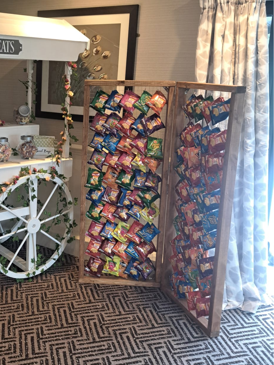 A freestanding wooden display stand with two panels filled with colourful packaged snack bags of crisps, photographed in an indoor room with patterned carpet, curtains, and a decorative sweets cart visible at left.