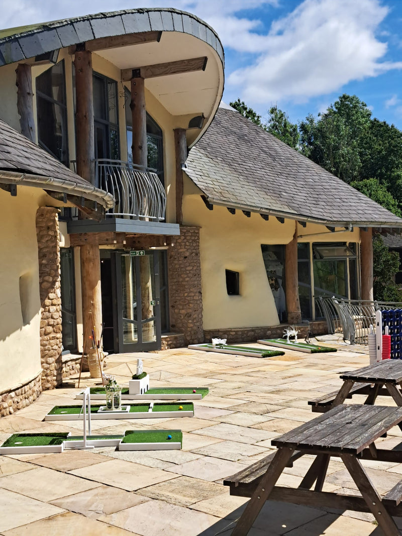 Outdoor patio scene in front of a modern lodge with curved roofs, stone and wood columns, and several mini golf greens set up on the paved terrace. Picnic tables are visible in the foreground, with trees and blue sky in the background. Forrest Hills, Lancaster University.