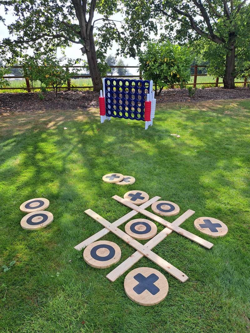 Outdoor giant games set up on green grass with a noughts & crosses game with wooden discs, plus a red, white, and blue Connect 4 game in the background near trees. The Aviary, Ormskirk.