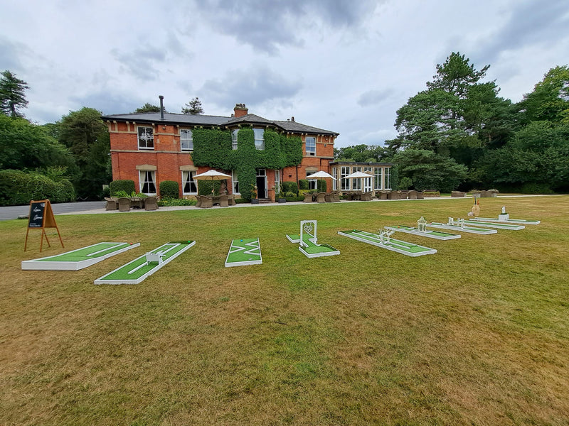 6 hole wedding mini golf courses set up on a large grassy lawn in front of a large red brick estate building with ivy, under a cloudy sky. Bartle Hall, Preston