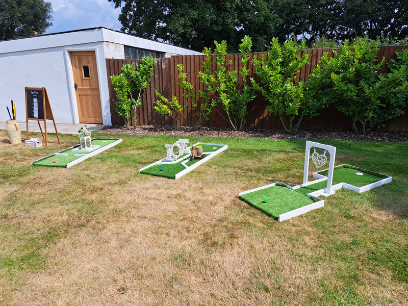 Outdoor white framed 3 hole mini golf setup on a dry grass lawn, with white decorative wedding themed props, a wooden fence, shrubs, and a small white building in the background. The Aviary, Ormskirk.