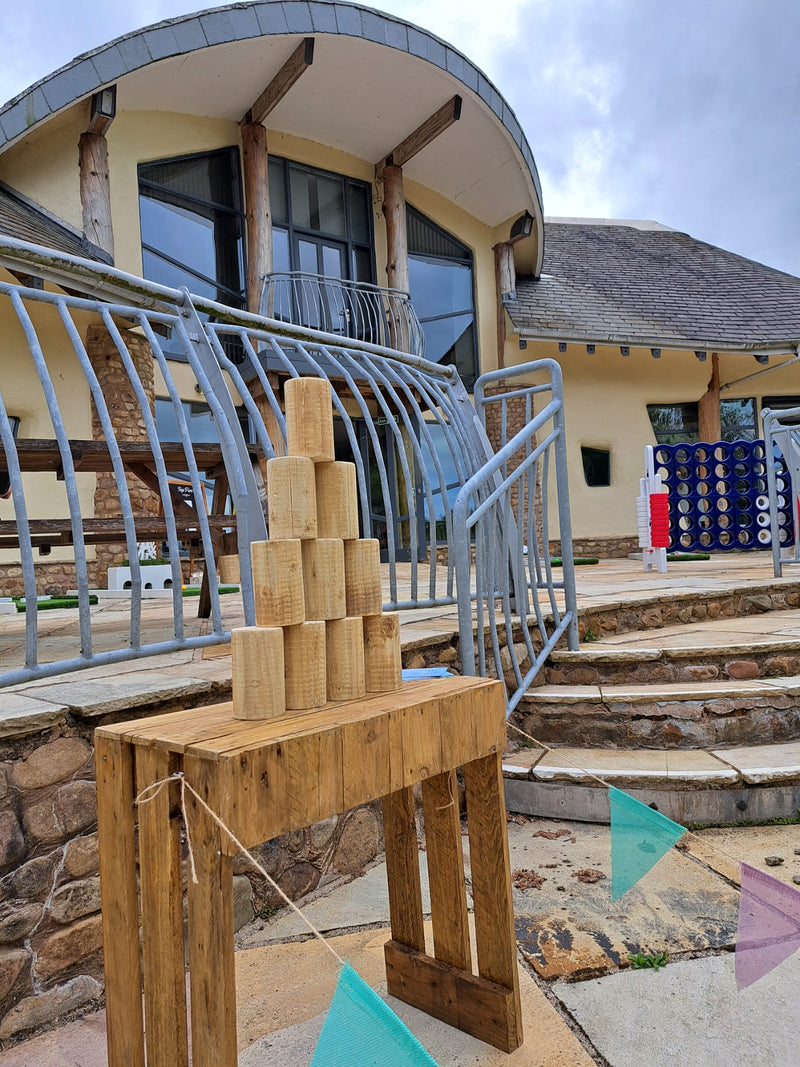 A rustic wooden tin can alley game  set outdoors in front of a modern building with stone walls and metal railings. The table holds a pyramid stack of natural wood blocks, with a 4 in a row game visible in the background. Forrest Hills, Lancaster University.