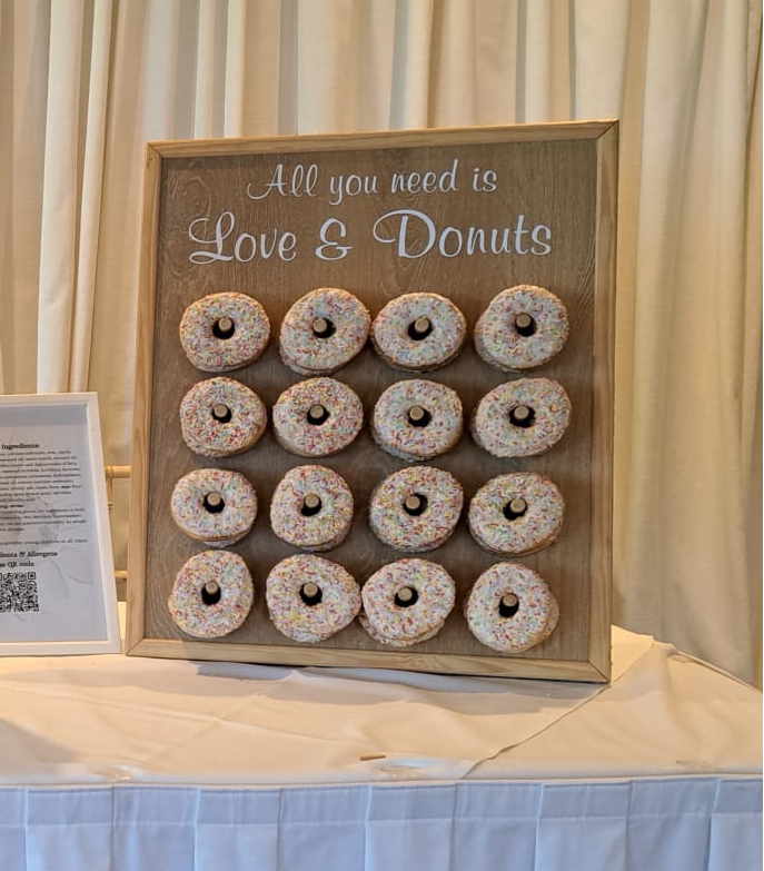 A rustic wooden sign with the phrase 'All you need is Love & Donuts' above a 4x4 grid of 16 frosted donuts with sprinkles, displayed on a draped tablecloth in front of cream curtains.