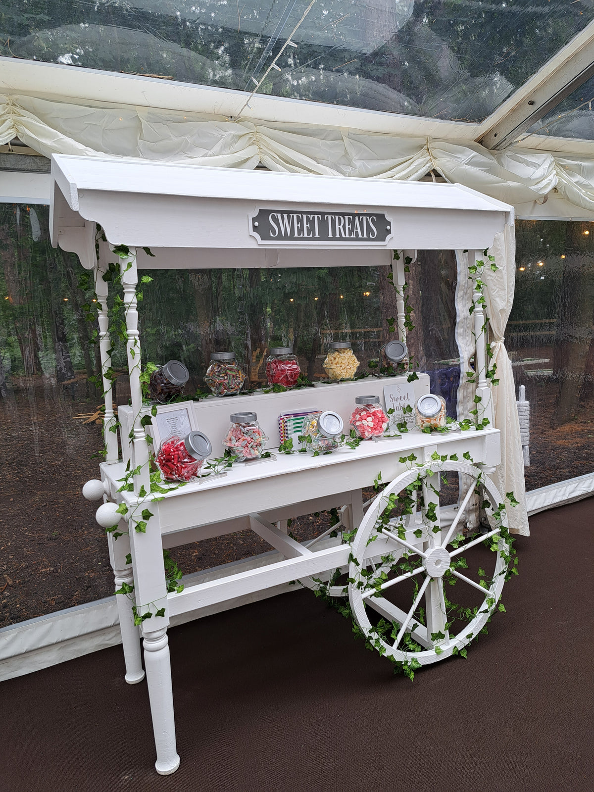 Decorative candy display with a white wheels and sweets inside a clear wedding marquee.