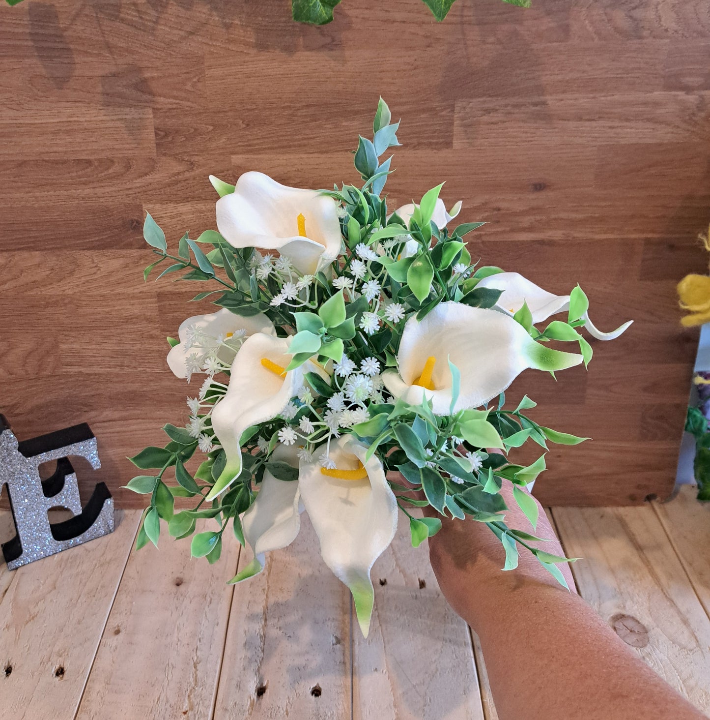 An artificial calla lily bouquet with green leaves and white baby's breath, held by a hand against a rustic wooden plank background.