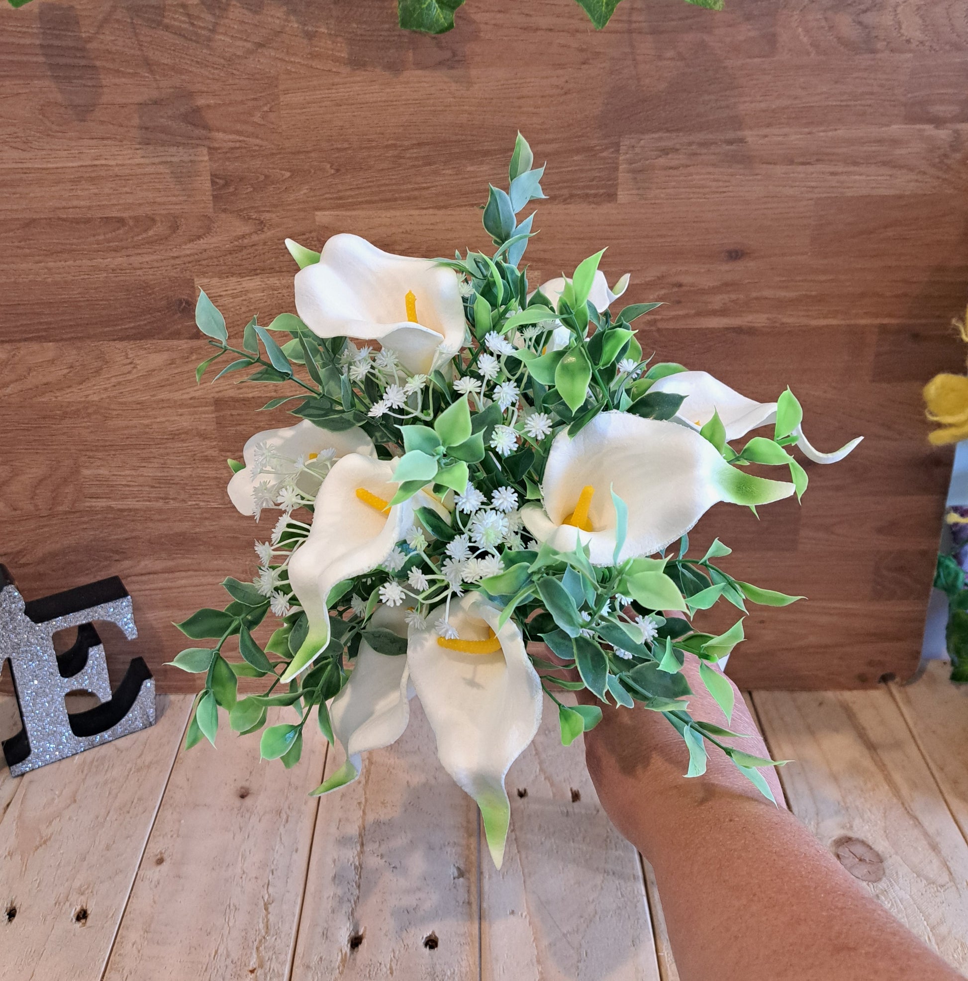 An artificial calla lily bouquet with green leaves and white baby's breath, held by a hand against a rustic wooden plank background.