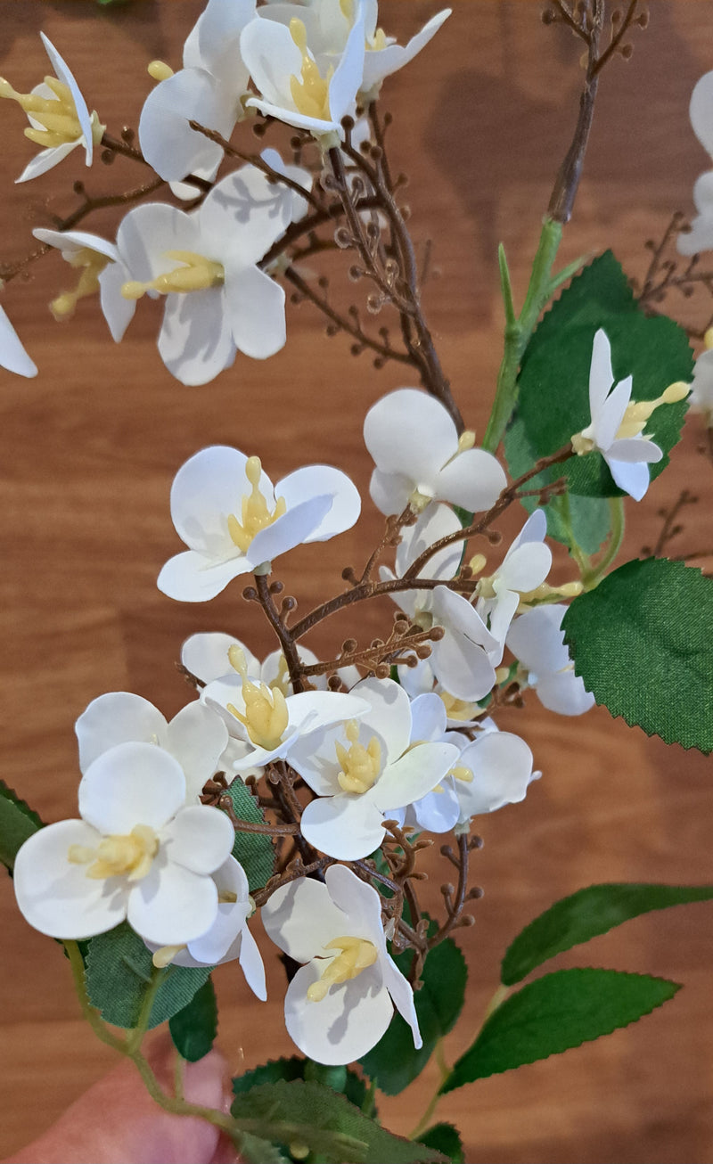 Artificial white blossom flowers on brown stems with green leaves set against a rustic background