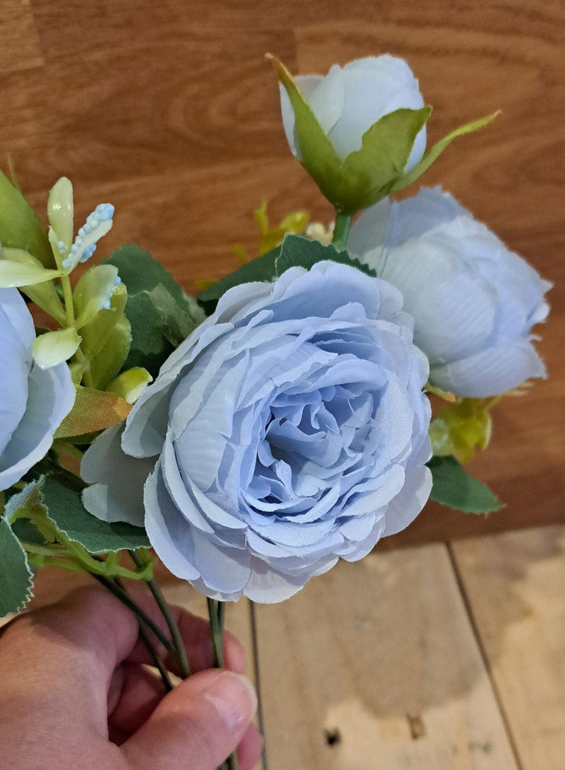A close-up of a hand holding a bouquet of artificial light blue peony roses and green foliage against a wooden background.