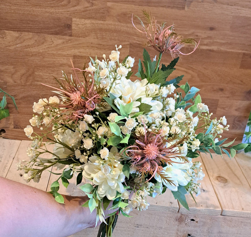 An artificial floral bouquet with cream flowers, green foliage, and dusty rose sea thistle, held by a person's arm against a wooden shelf background.