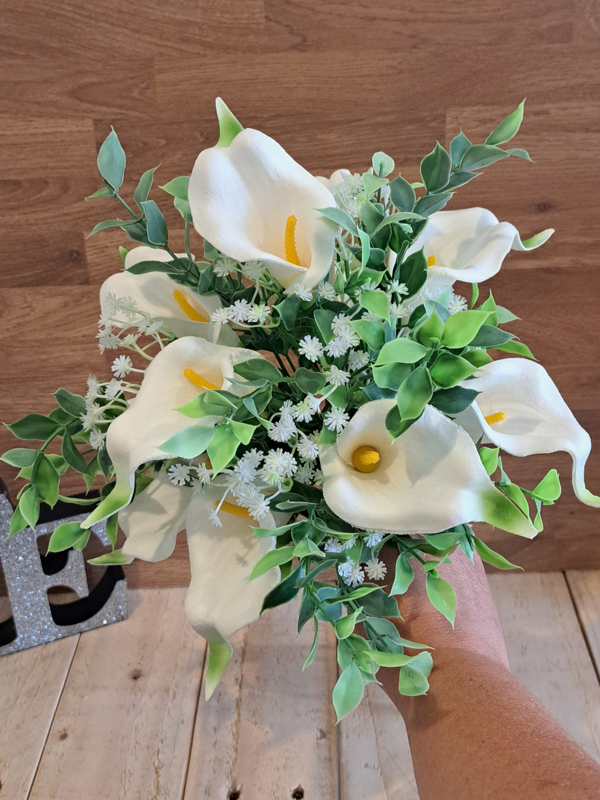 A bouquet of artificial white calla lilies with green leaves and small white filler flowers, held by a hand against a wooden background.