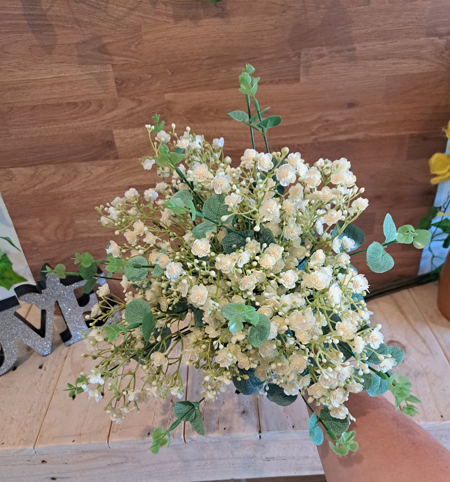 An artificial floral arrangement featuring ivory-coloured baby's breath flowers with green leaves, held by a hand against a wooden wall and floor background.