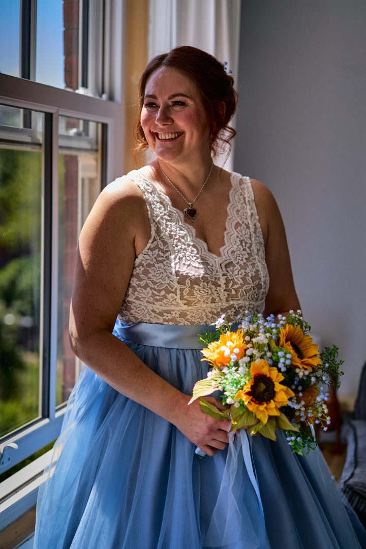 A bride in a white lace top and blue skirt holding a bouquet of sunflowers by a window.