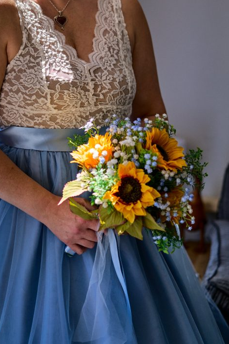 A bride wearing a lace top and blue skirt holding a bouquet of sunflowers and other flowers.