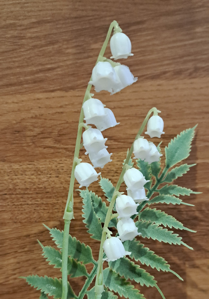 An artificial lily of the valley flower stem with green leaves, photographed against a brown wood-grain background.