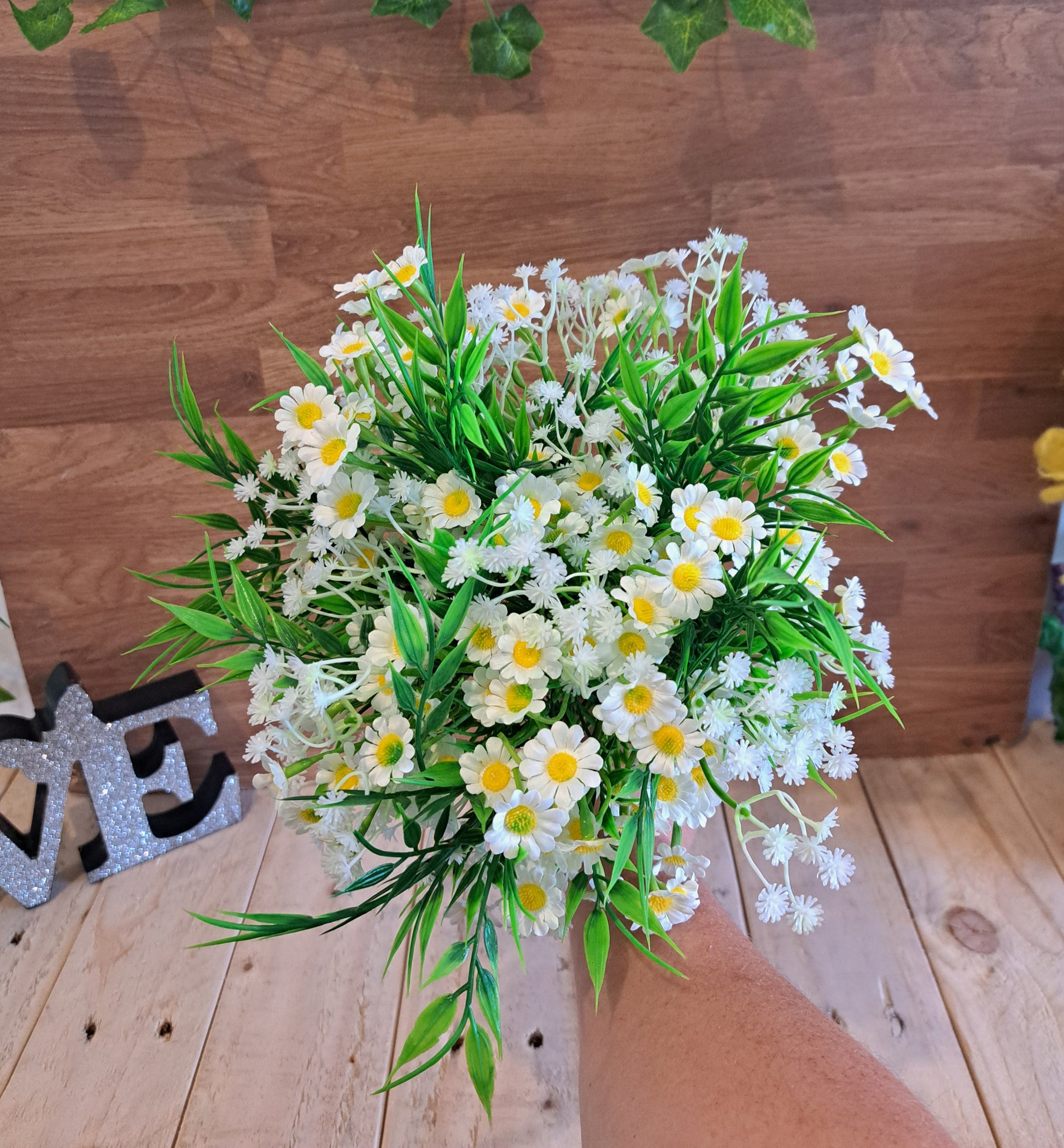 A bouquet of artificial white daisy flowers with yellow centres, baby breath and green leaves, held by a person's arm against a wooden wall and floor background.