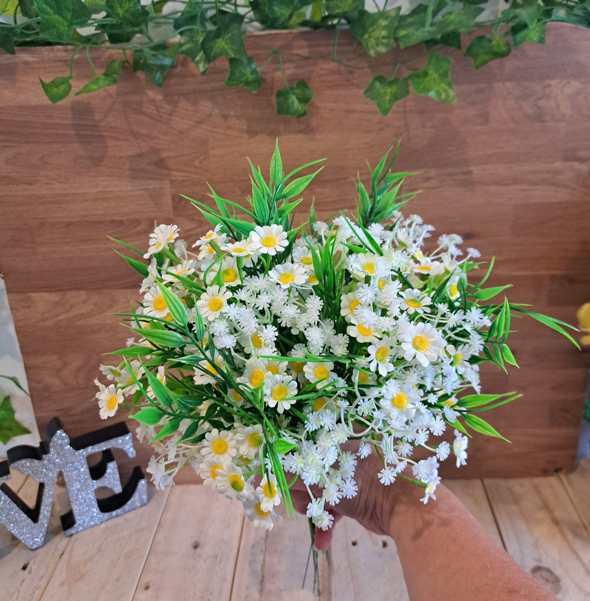 A hand holding a bouquet of artificial white daisies with yellow centers and green leaves, set against a wooden wall and floor backdrop.