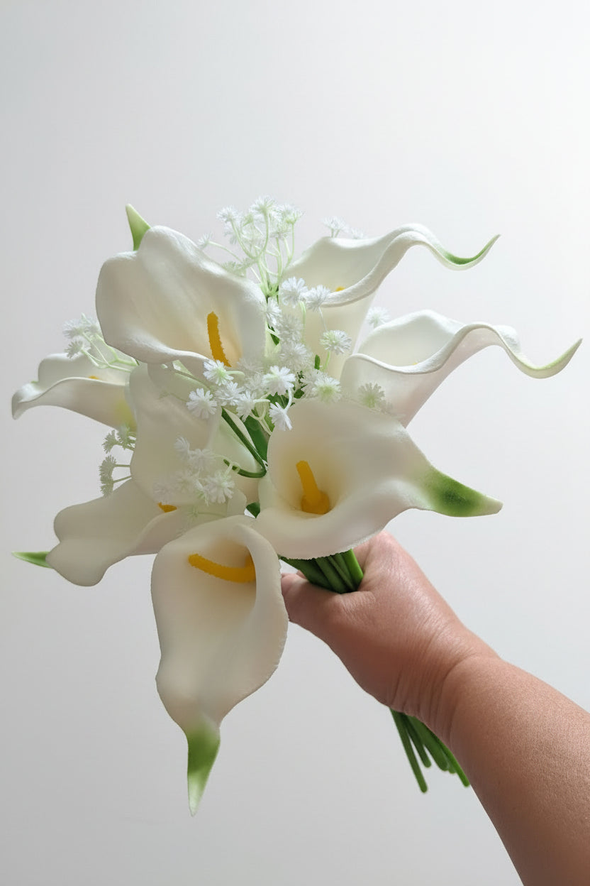 A bouquet of artificial white calla lilies with yellow centres and small white filler flowers, held in a person's hand on a white background.