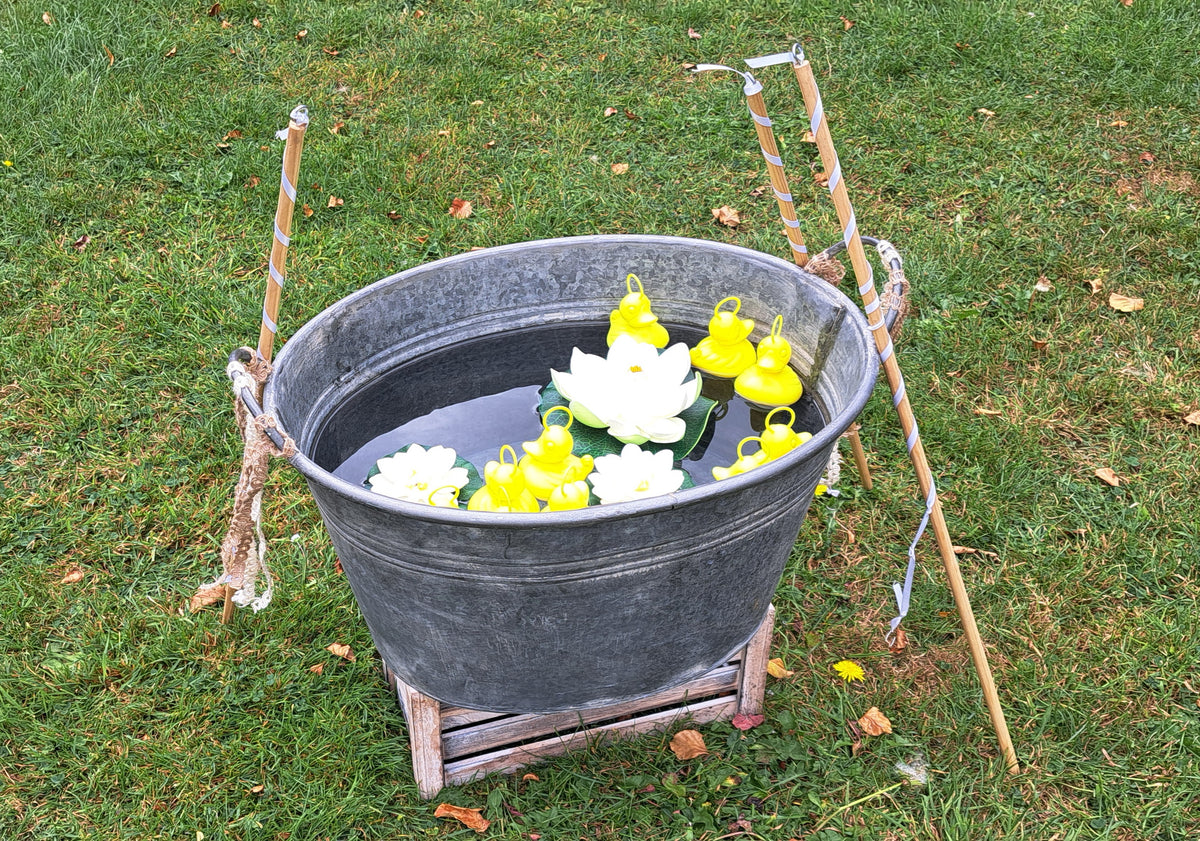 A galvanized metal tub filled with water, yellow hook a ducks and floating lotus-shaped flowers, set on a small wooden stand outdoors on a grassy lawn.