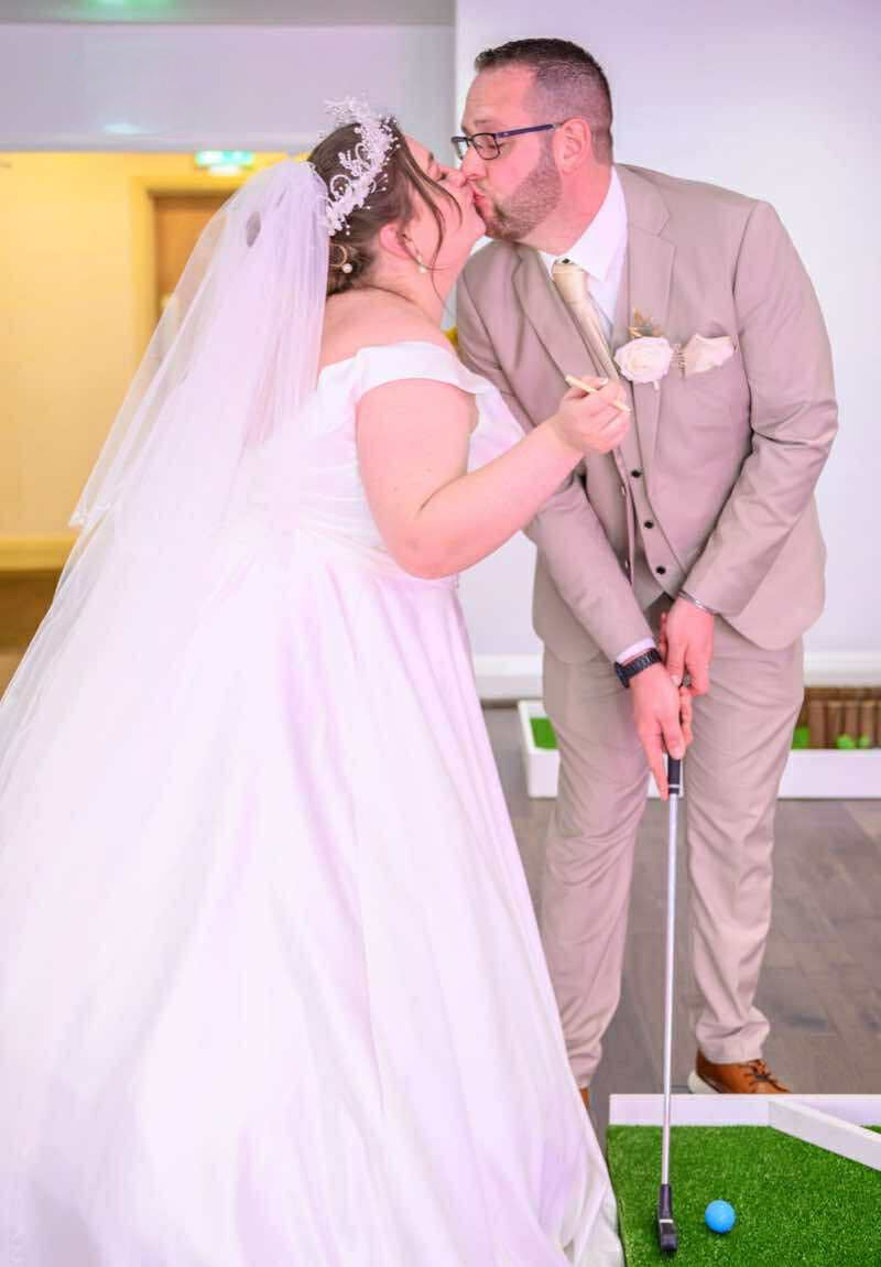 A bride in a white wedding dress and veil and a groom in a light beige suit playfully kiss while standing on a mini golf course. The Carousel Hotel, Blackpool.