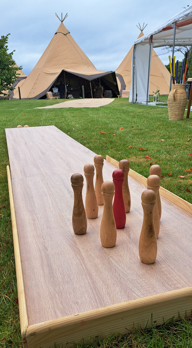 A wooden outdoor pinball game set with natural wood pins and a red accent pin on a light wood board, placed on a grassy lawn with teepees in the background. Parr Hall, Eccleston