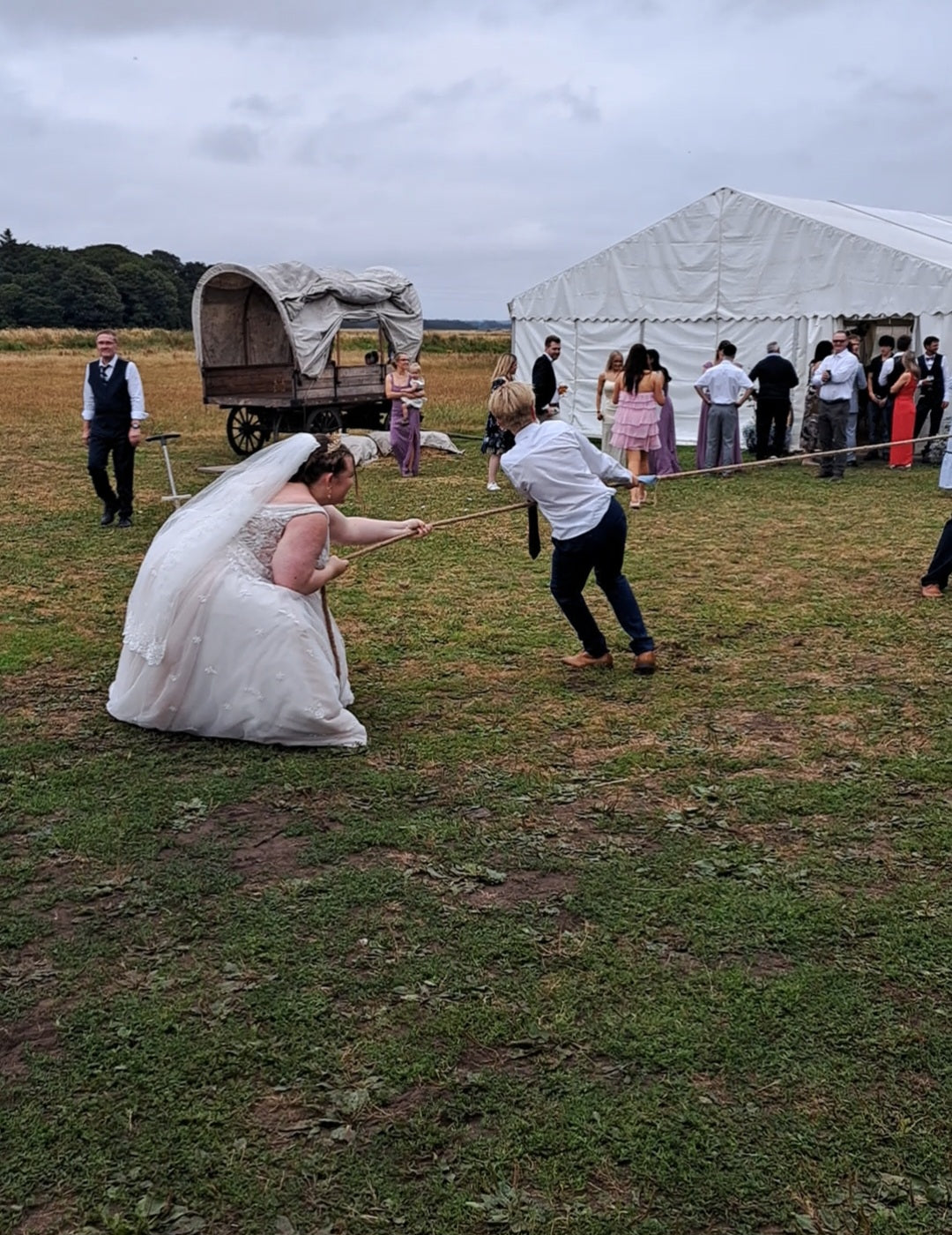 A bride in a white wedding gown and veil and a man in a white shirt and dark trousers pull on a rope outdoors on grass, with a white event tent, a covered carriage, and several guests in the background.