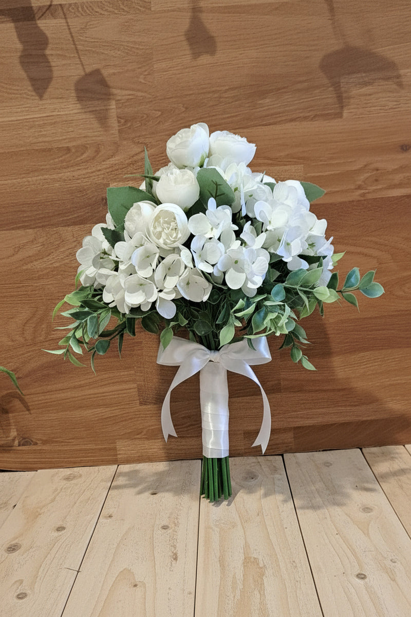 A bouquet of white faux roses and hydrangeas with green leaves, set against a wooden wall and shelf background.