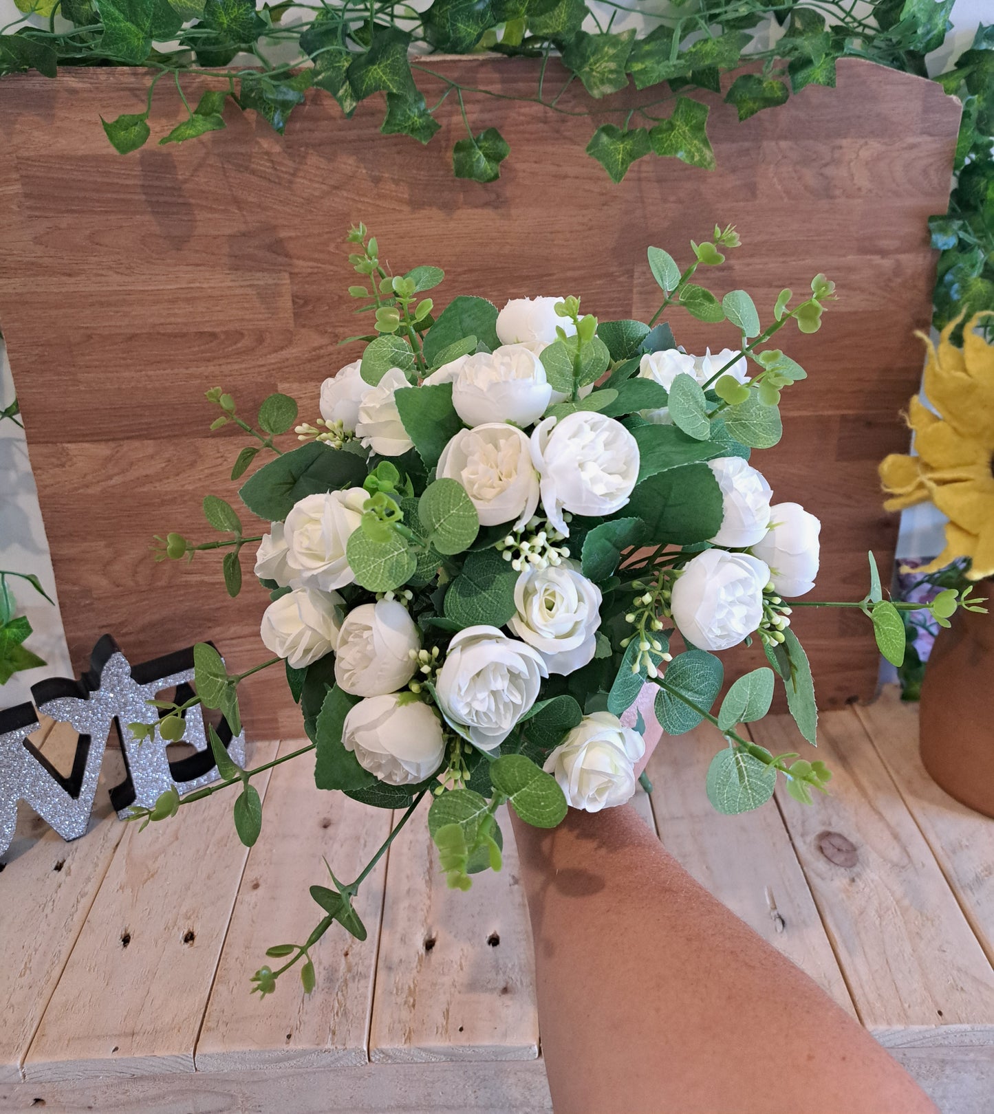 An artificial flower bouquet with white roses and green eucalyptus leaves, held by a person's hand in front of a wooden backdrop.