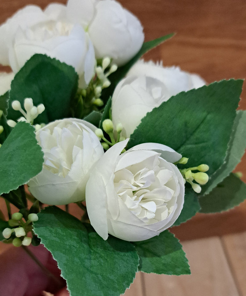 White faux peony artificial flower bouquet with green leaves and small buds, shown close up against a wooden background.