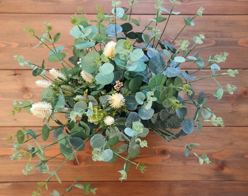An artificial floral arrangement with green eucalyptus leaves and cream-colored flowers, held by a person's hand in front of a wooden background.