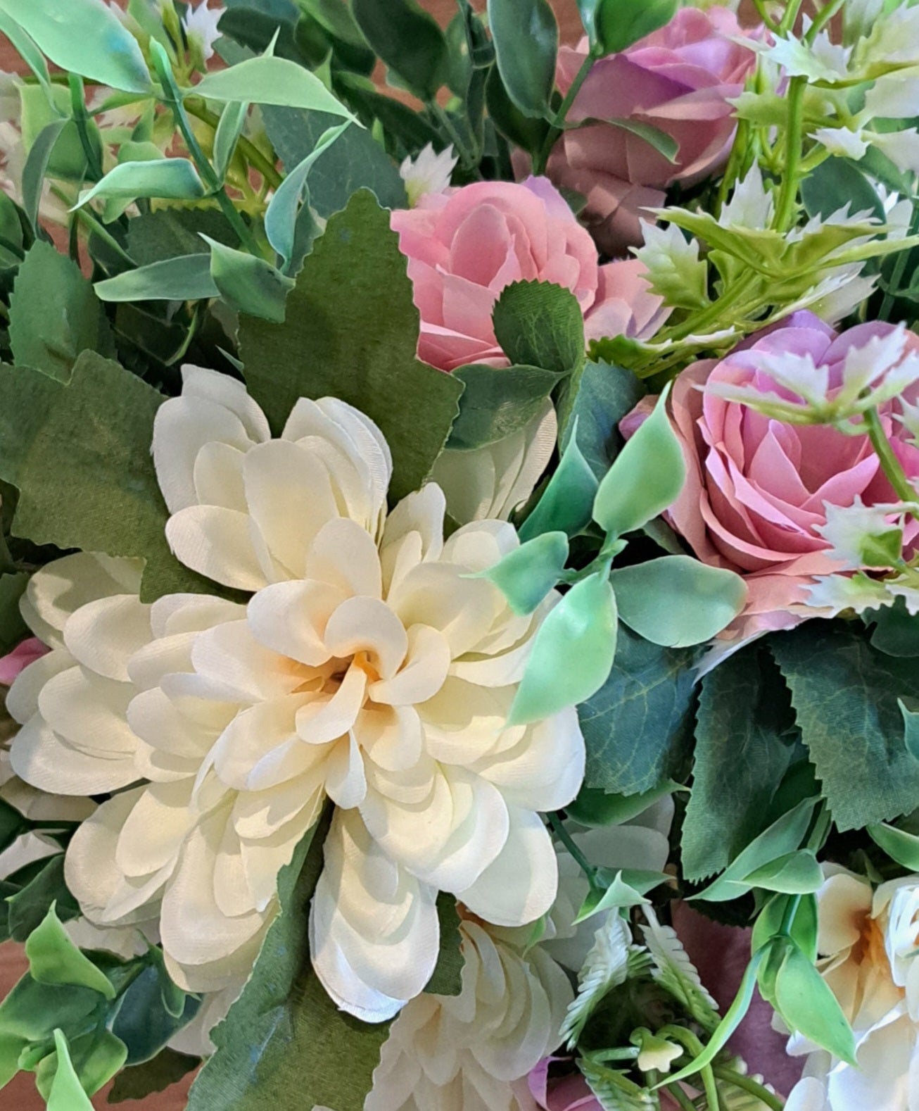 A enlarged view of a bouquet of artificial flowers, including pink roses and cream dahlia with green leaves, set against a rustic wooden plank background.
