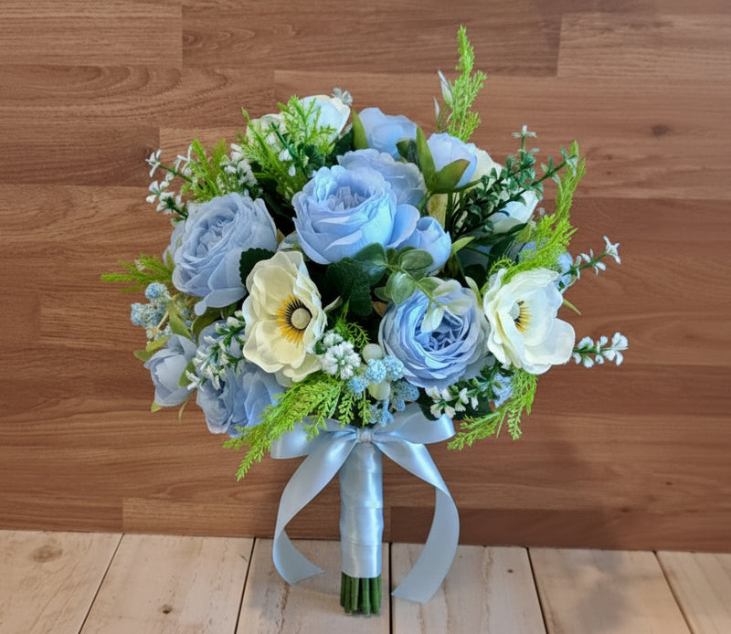 A bouquet of artificial flowers, featuring light blue roses, pale ivory anemones, and green foliage, set against a rustic wooden plank background.