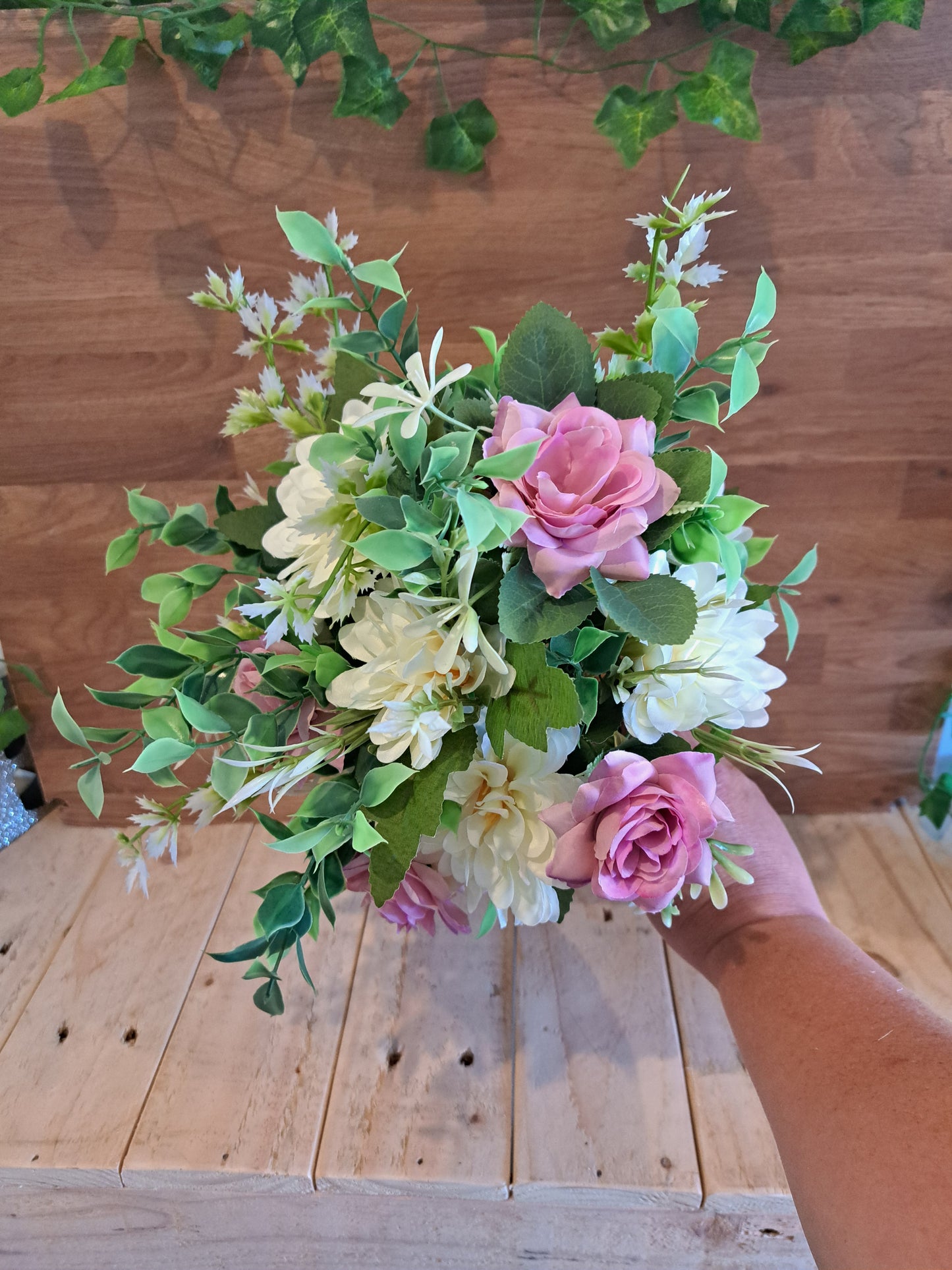 A hand holding a bouquet of artificial flowers, including pink and cream roses with green leaves, set against a rustic wooden plank background.