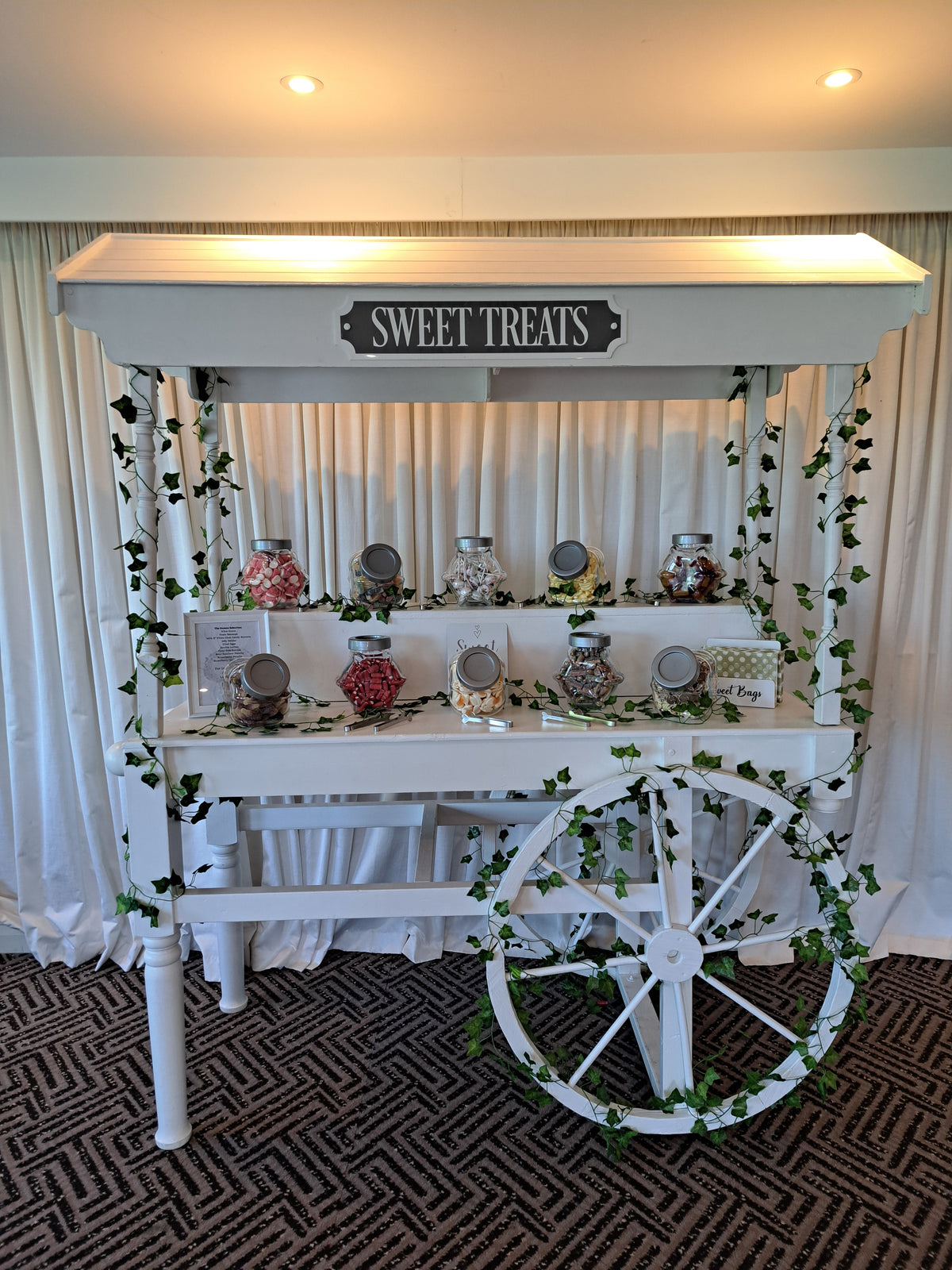 A white decorative cart styled as a sweet treats stand, with a canopy labelled 'SWEET TREATS,' glass jars filled with candies, green ivy garlands, and a large spoked wheel on a patterned carpet in front of white curtains. Wrightington Hotel & Spa, Wigan.