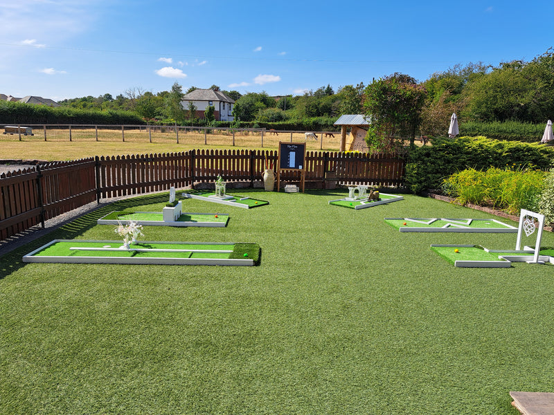 An outdoor 6 hole wedding mini golf course setup on a green artificial turf, with multiple green putting surfaces, a dark wooden fence, trees, and a blue sky in the background. The Barn at Yew Tree Farm, Halewood.