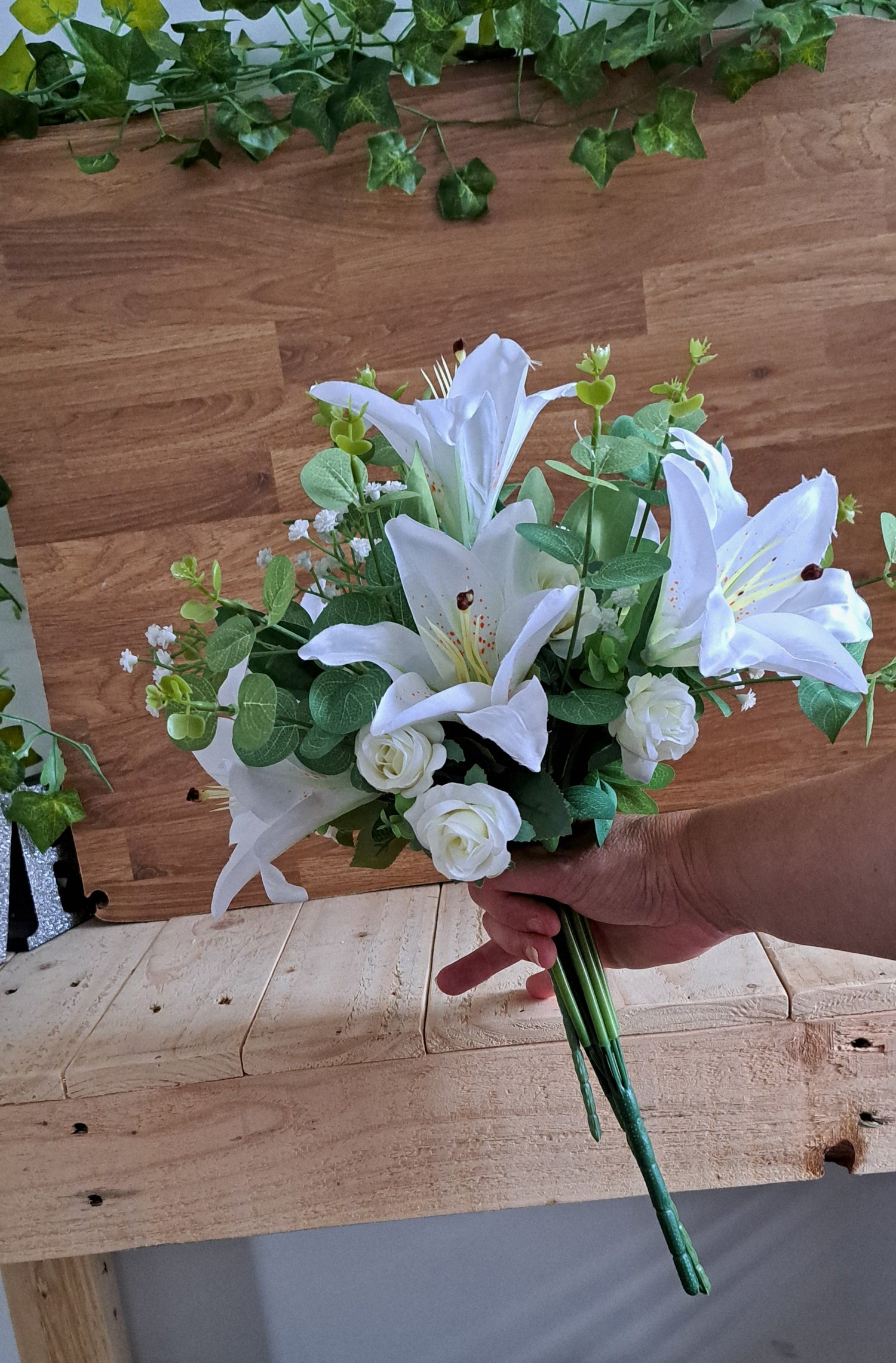 A bouquet of artificial white lilies and roses with green foliage, held by a person's hand in front of a wooden shelf and wall.