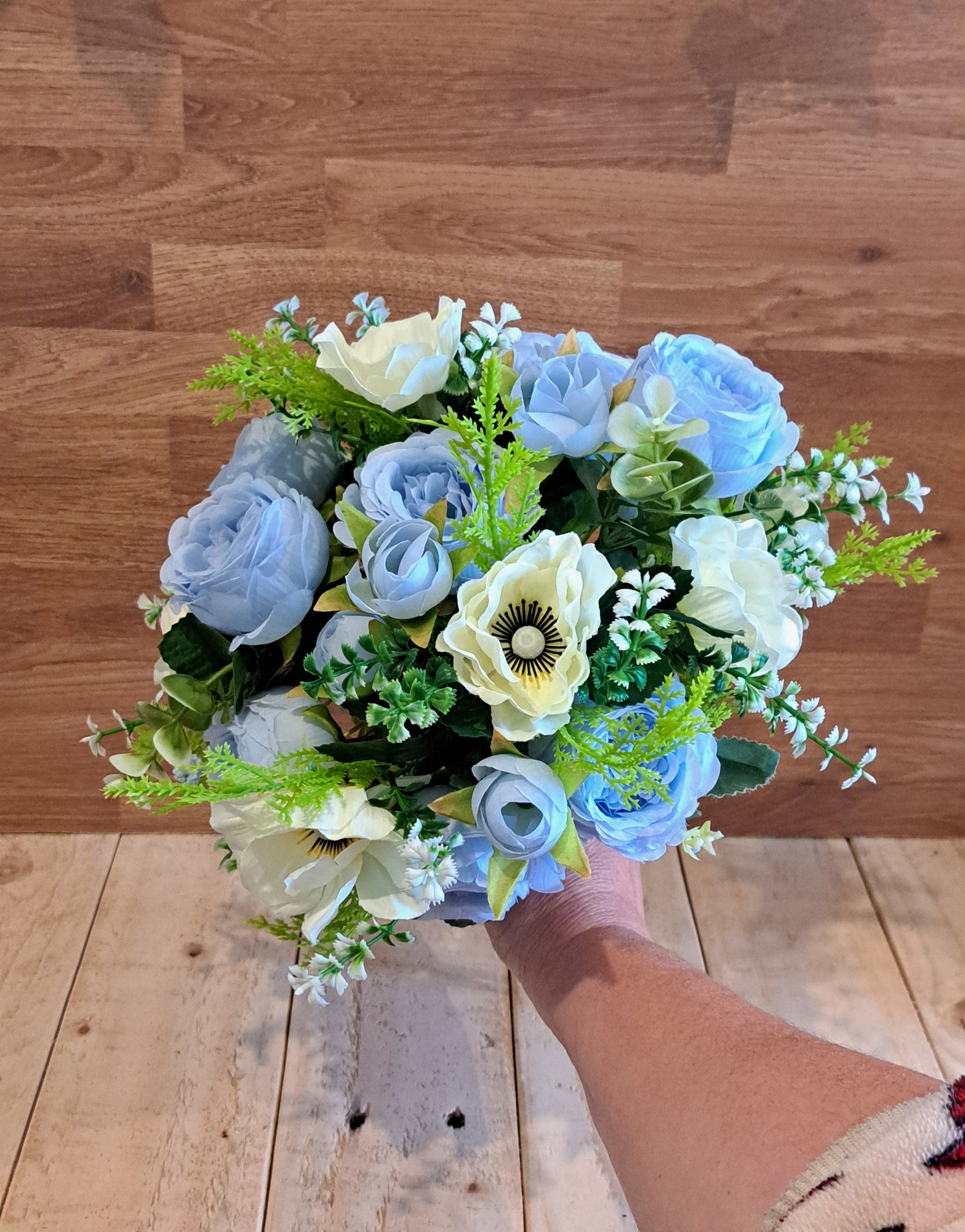 A bouquet of artificial flowers in light blue, cream, and green, held by a person's hand in front of a wooden wall and bench.