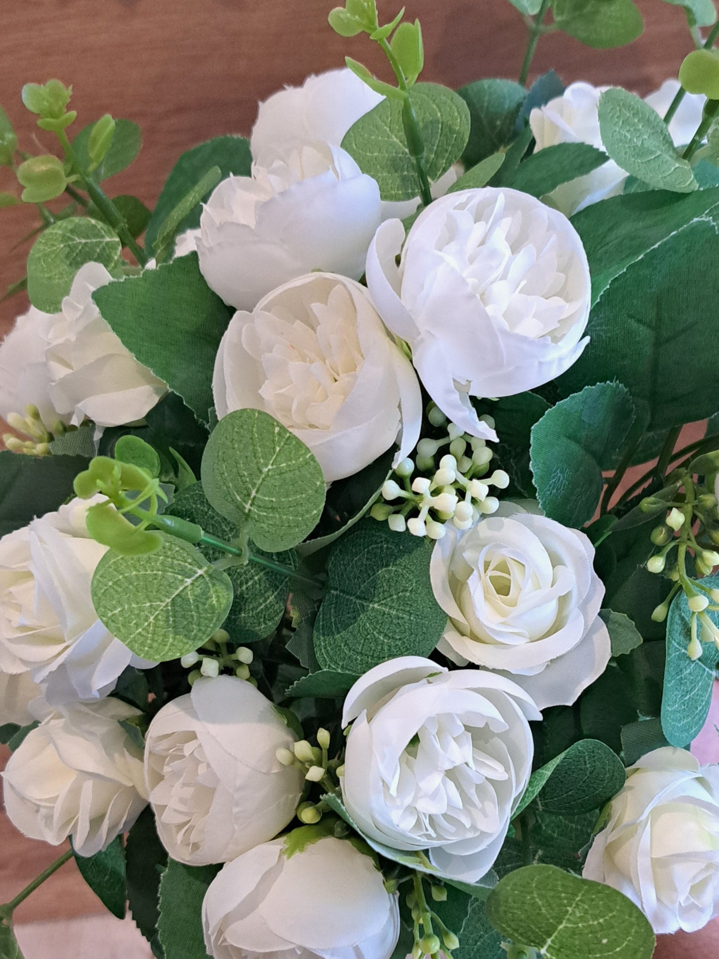 A close-up of an artificial floral arrangement featuring white roses and green leaves, photographed against a warm brown wooden background.