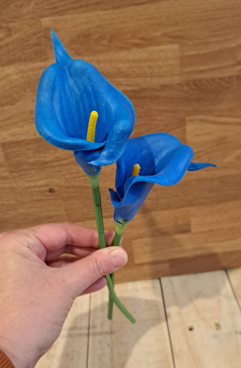 A hand holding two artificial blue calla lily flowers with green stems and yellow centers, set against a wooden background.
