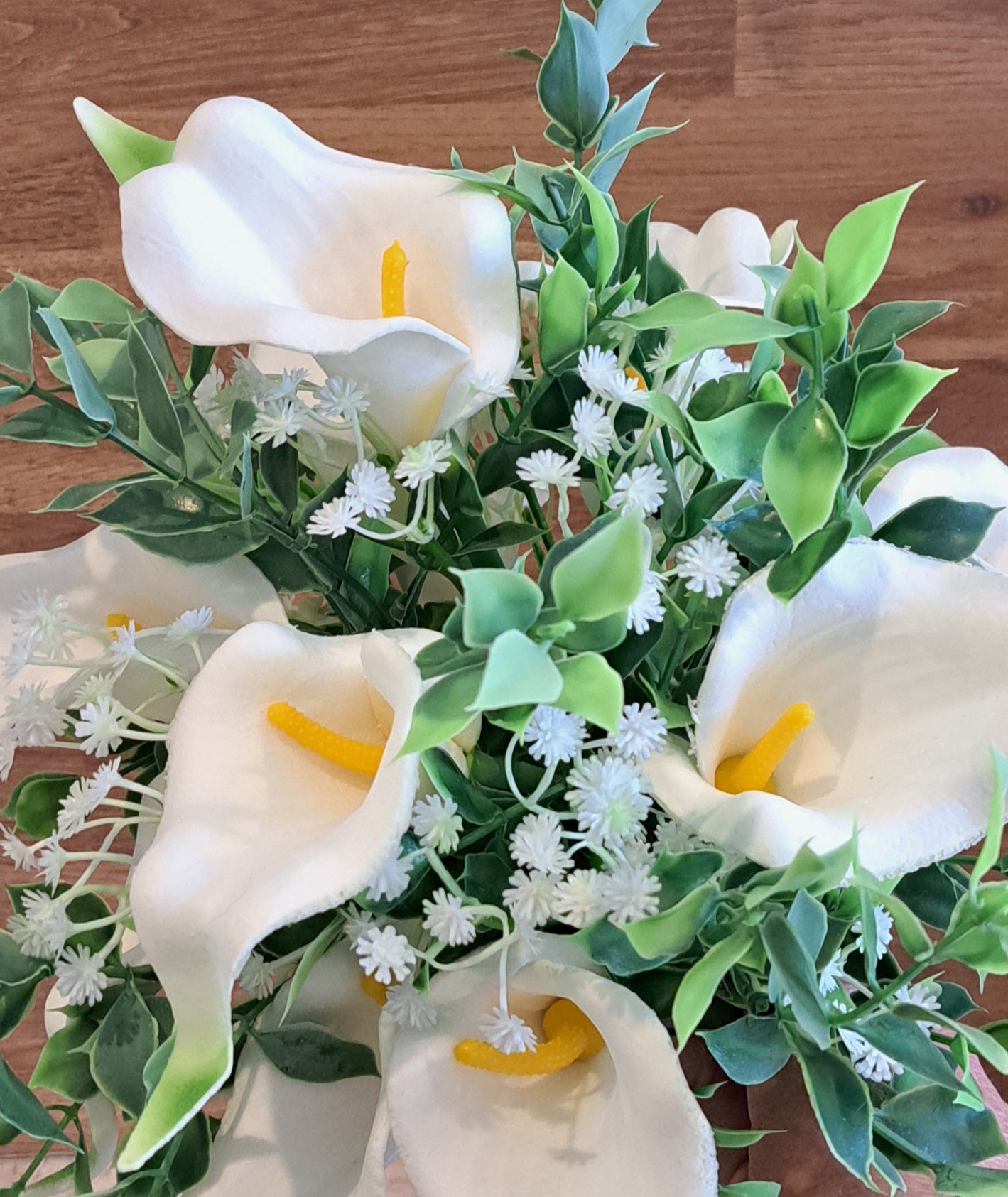 A close up  view of an artificial calla lily bouquet with green leaves and white baby's breath, held by a hand against a rustic wooden plank background.