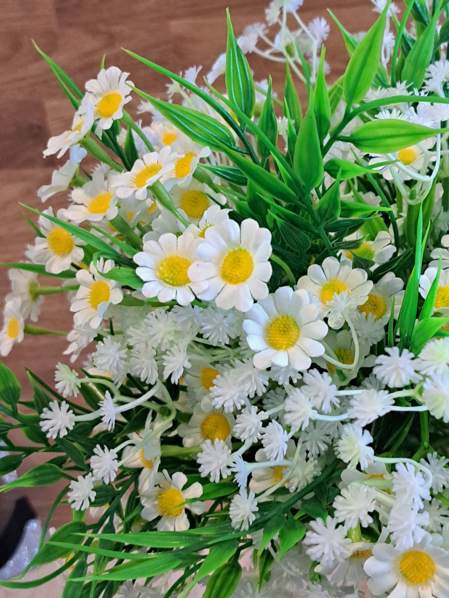 A close up view of an artificial daisy bouquet with green leaves and white baby's breath, held by a hand against a rustic wooden plank background.